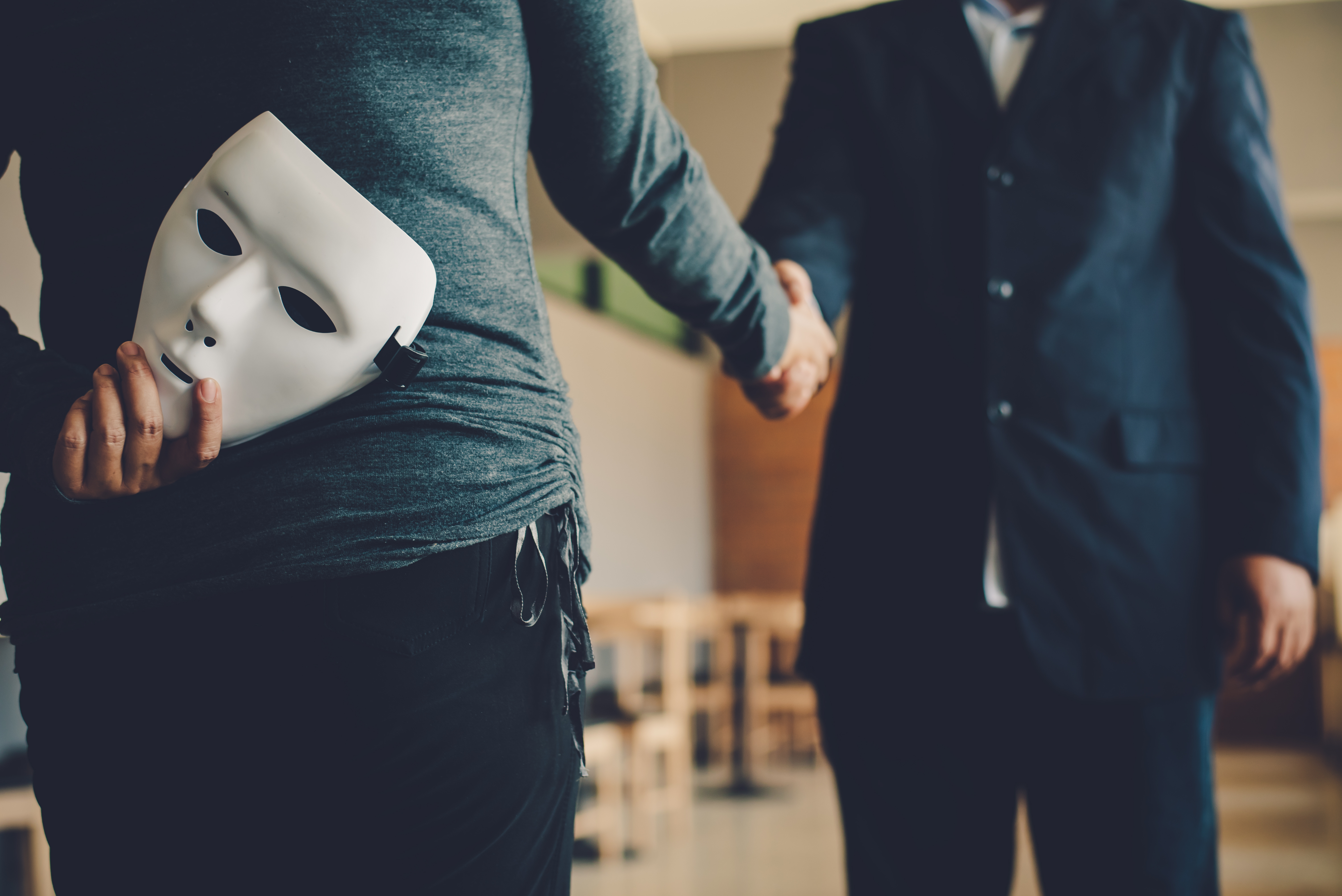 An image of a man holding out his hand to shake, but he is holding a white mask behind his back, cueing that he is untrustworthy — For an expereinced Utah theft by deception defense attorney, contact Branson West Law.